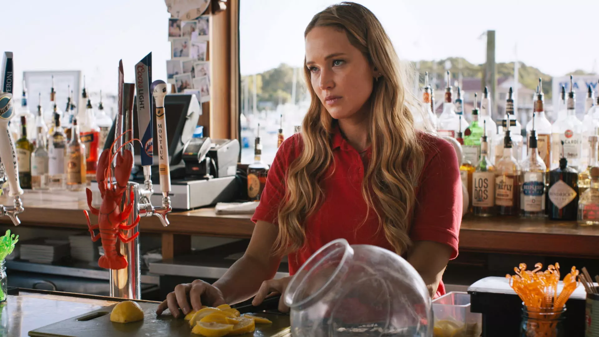 جنیفر لارنس با لباس فرم قرمز ایستاده پشت میز یک بار ساحلی در حال بریدن لیمو در نمایی از فیلم بدون احساسات جدی به کارگردانی جین استوپینتسکی