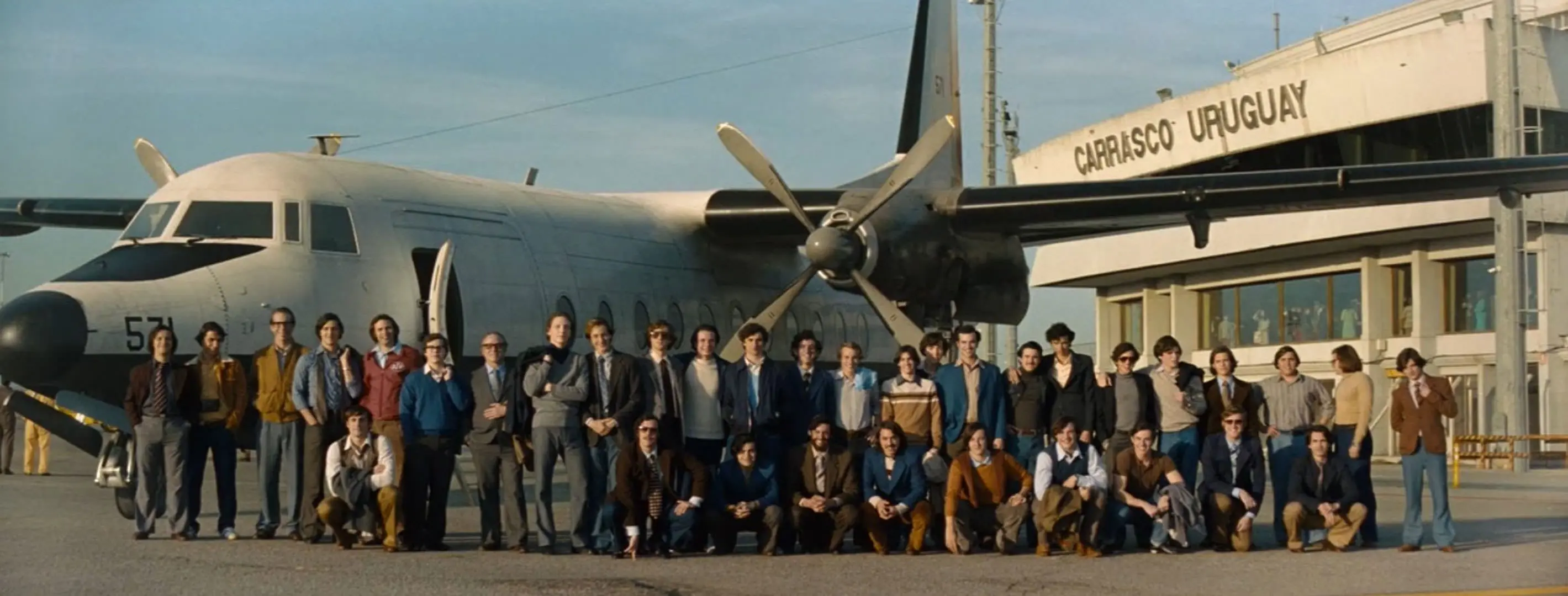 پرواز ۵۷۱ نیروی هوایی اروگوئه با مسافرانی که در فرودگاه مقابل هواپیما ایستاده‌اند و عکسی دسته‌جمعی را به یادگاری می‌گیرند در نمایی از فیلم انجمن برف به کارگردانی جی ای بایونا
